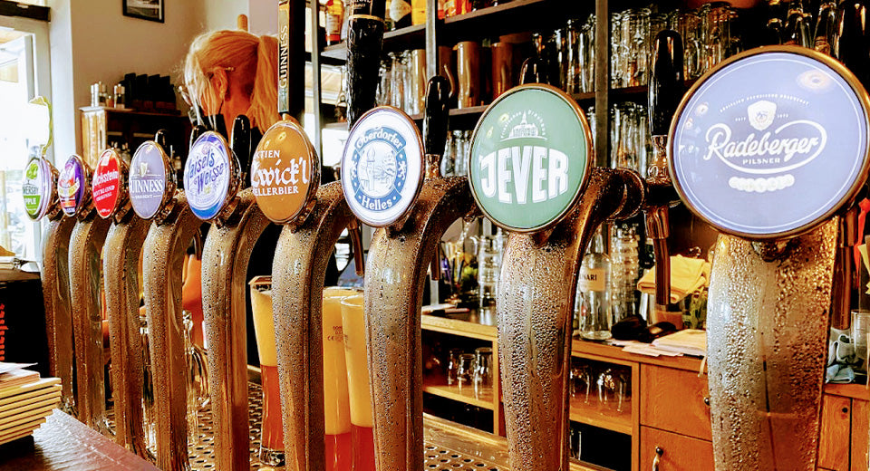 Zapfhähne verschiedener Brauerein wie Radeberger, Jever, Zwick'l und Guinnes nebeneinander, im Hintergrund eine Bar.