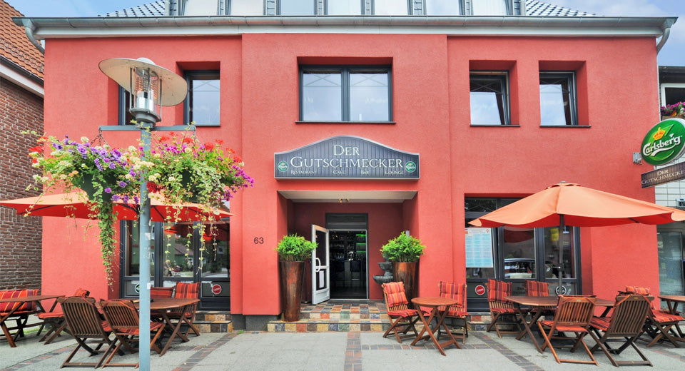 Eingangsbereich eines roten Gebäudes, davor Tische und das Logo des Restaurants "Der Gutschmecker".