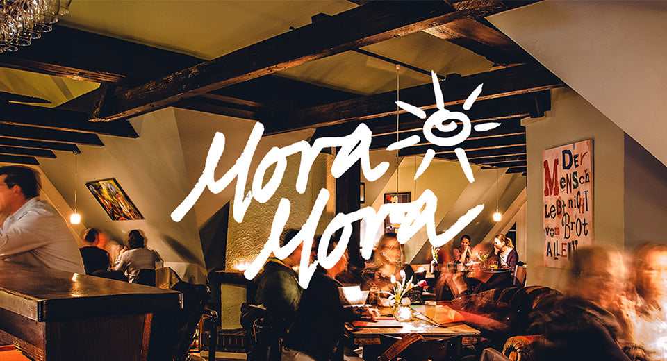 Eine Resturant Szene in einem Restaurant mit großen Balken, hell hell beleuchtet ohne Fenster. Darauf der Schriftzug "Mora Mora" mit einer Sonne.