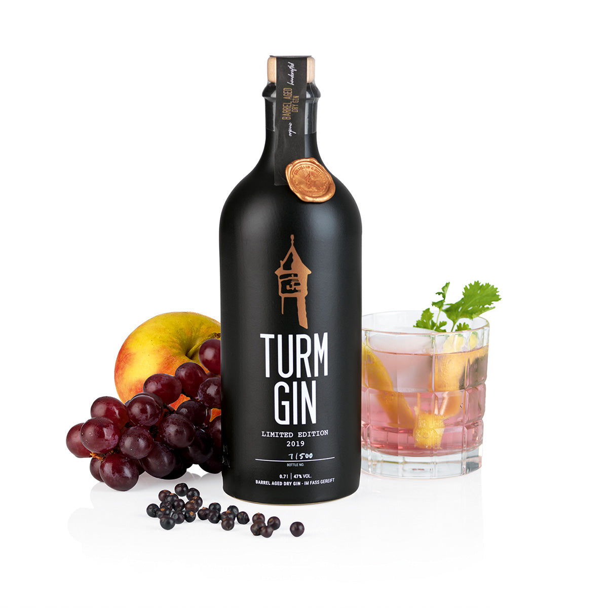 Die schwarze Flasche der TURM GIN Limited Edition 2019, daneben Äpfel, Trauben und ein fertiger Gin-Cocktail vor weißem Hintergrund.