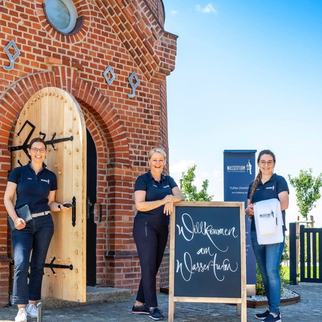 Drei Frauen stehen vor der Holz-Eingangstür des Wasserturm in Bad Segeberg, davor ein Schild "Willkommen am Wasserturm".