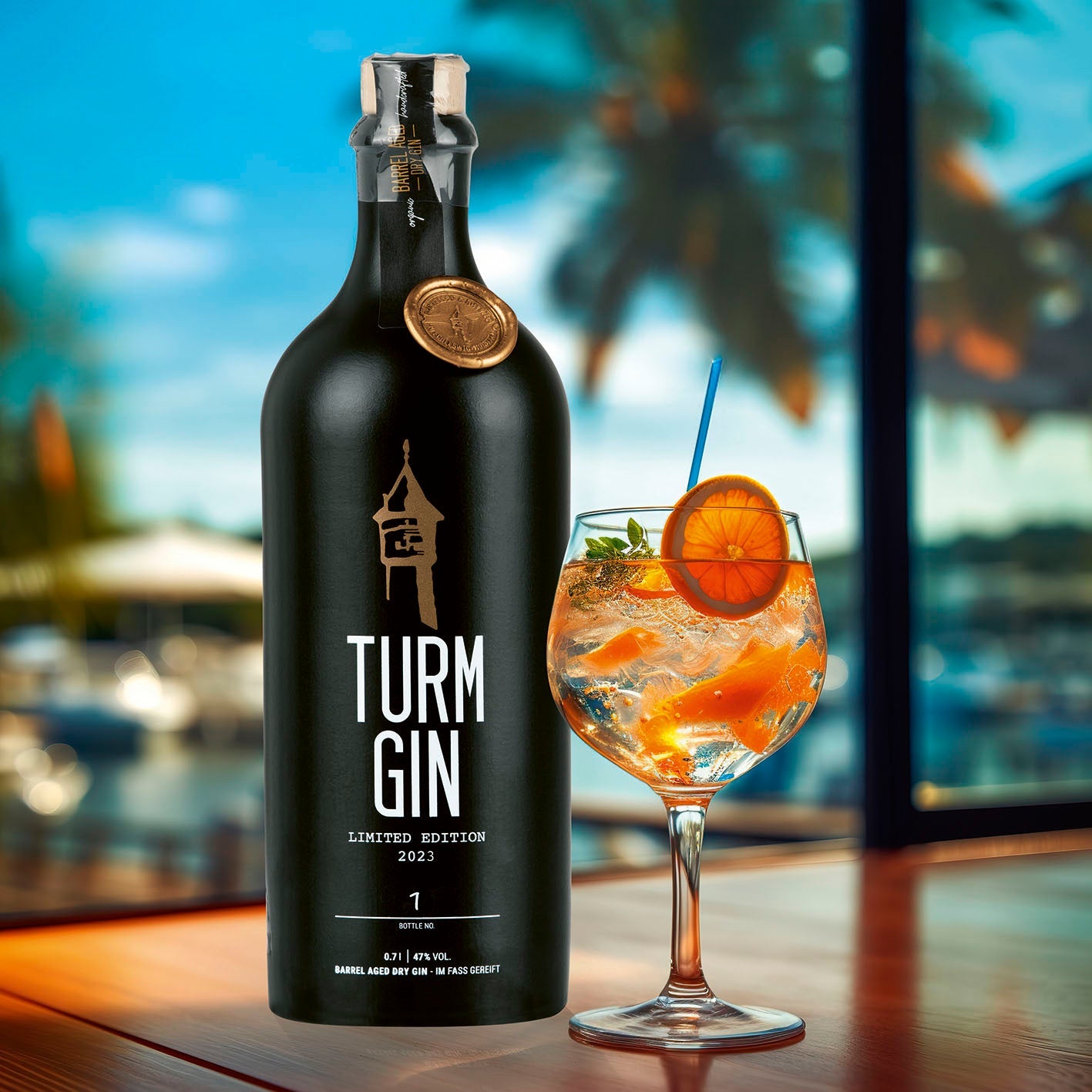 Schwarze Flasche TURM GIN Limited Edition 2023 neben einem Cocktailglas mit Orangen auf einem Tisch, im Hintergrund eine Poollandschaft.