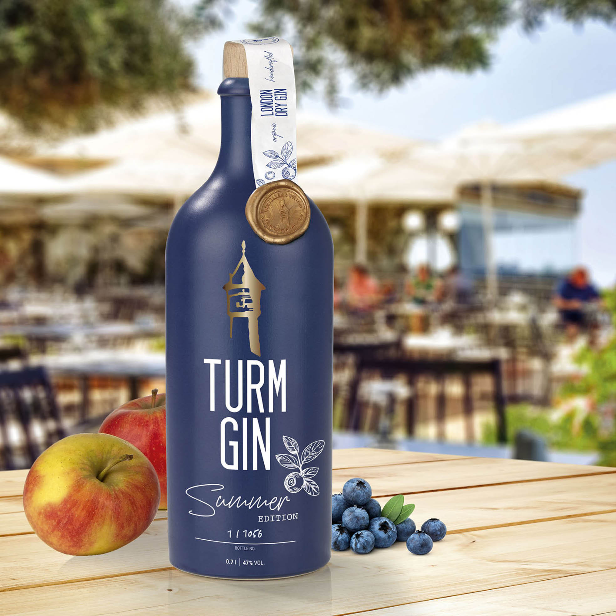 Blaue Flasche der TURM GIN Limited Summer Edition mit Turmsiegel, daneben Äpfel und Blaubeeren, im Hintergrund eine Freiluft Barszene.