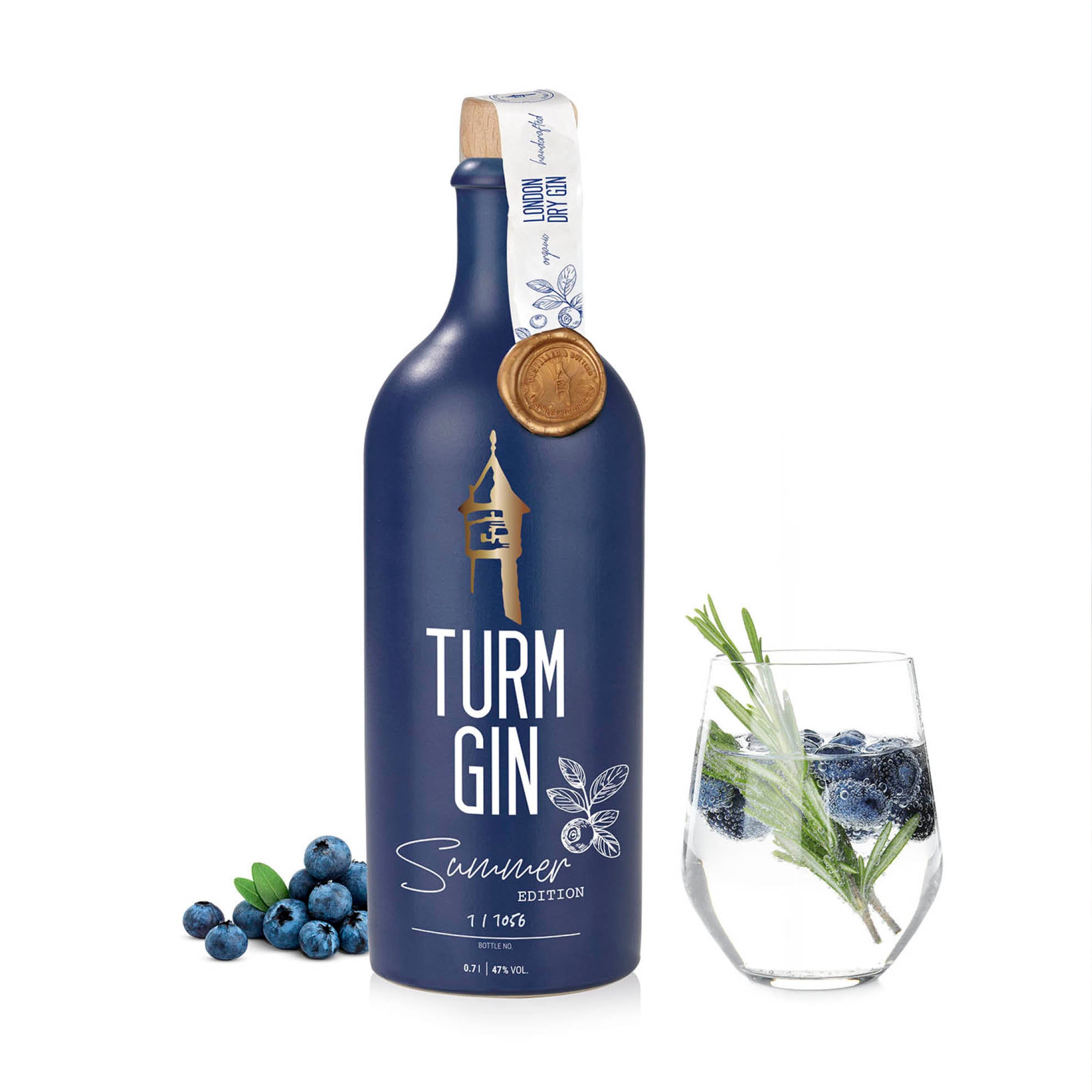 Blaue Flasche der TURM GIN Limited Summer Edition mit Turmsiegel, daneben Blaubeeren und ein Gin mit Blaubeeren vor weißem Hintergrund.