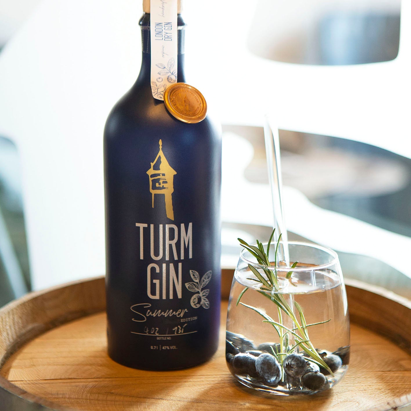 Blaue Flasche der TURM GIN Limited Summer Edition, daneben ein Gin mit Blaubeeren auf einem Holzfass.