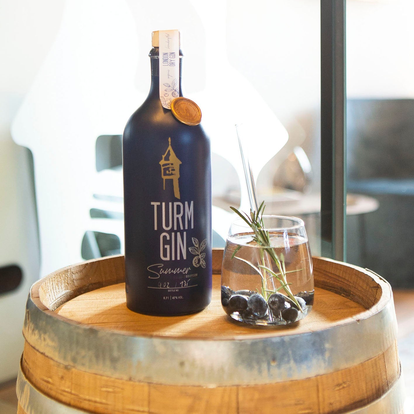 Blaue Flasche der TURM GIN Limited Summer Edition, daneben ein Gin mit Blaubeeren auf einem Holzfass.