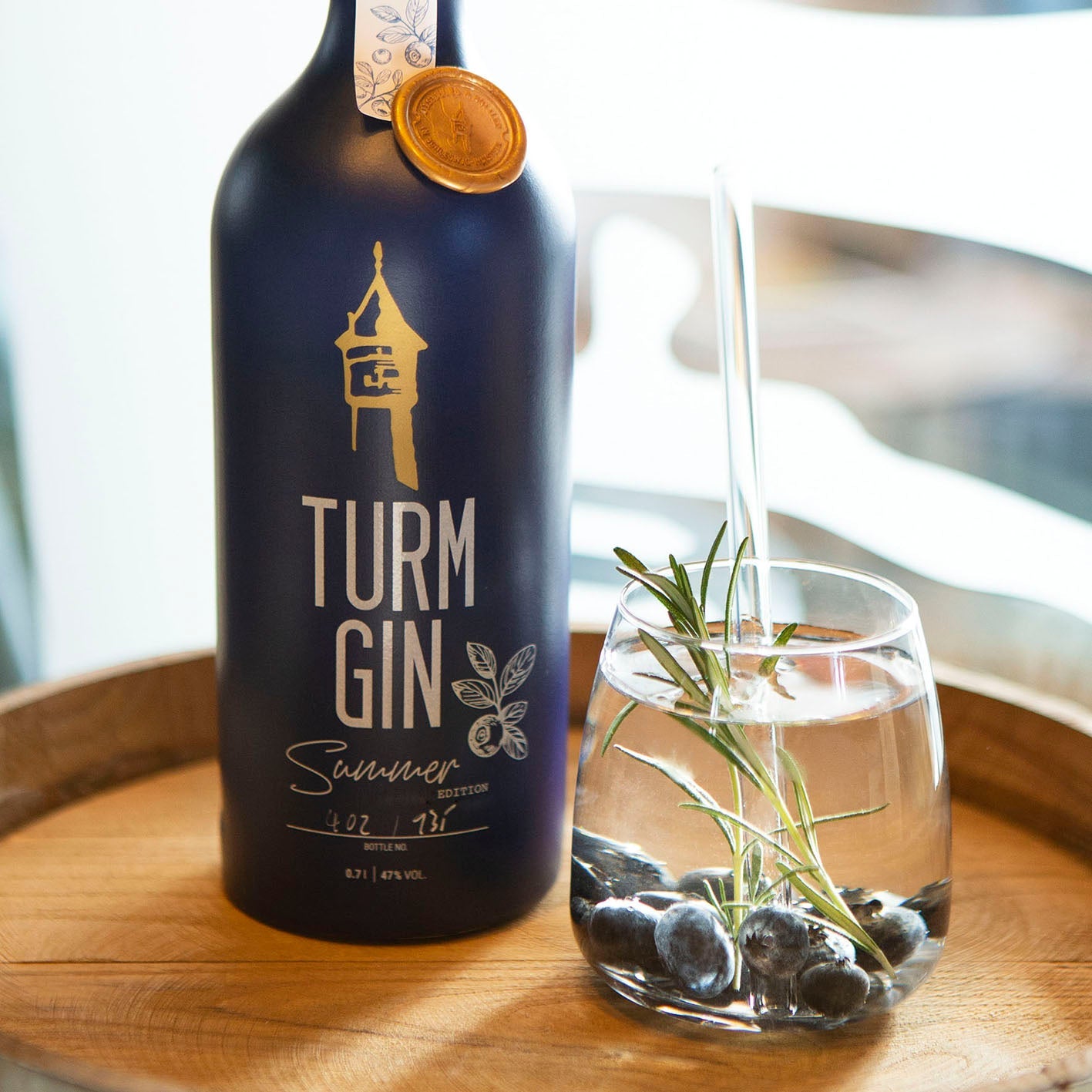 Blaue Flasche der TURM GIN Limited Summer Edition, daneben ein Gin mit Blaubeeren auf einem Holzfass.