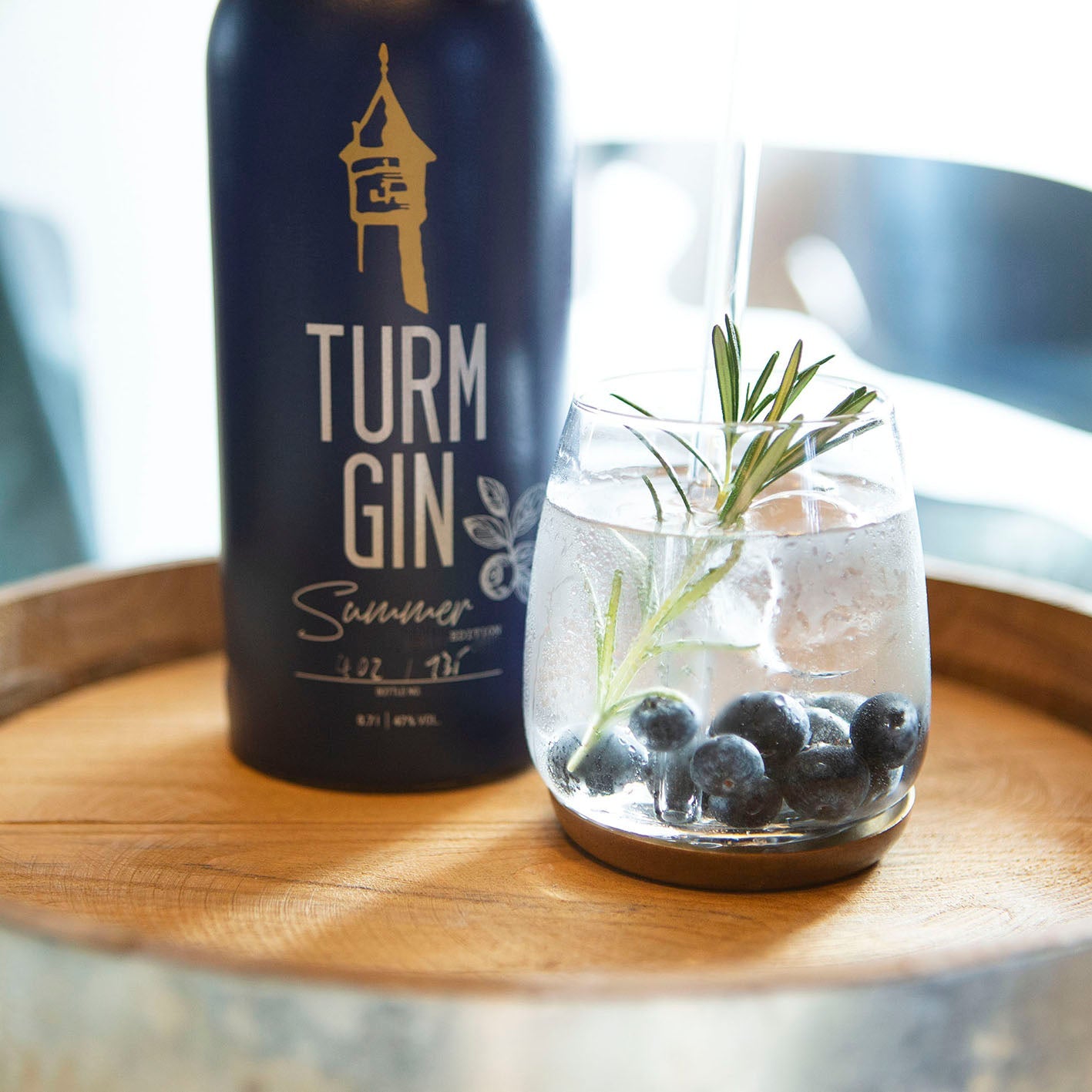 Gin mit Blaubeeren auf einem Tablett, dahinter die blaue Flasche der TURM GIN Summer Edition.