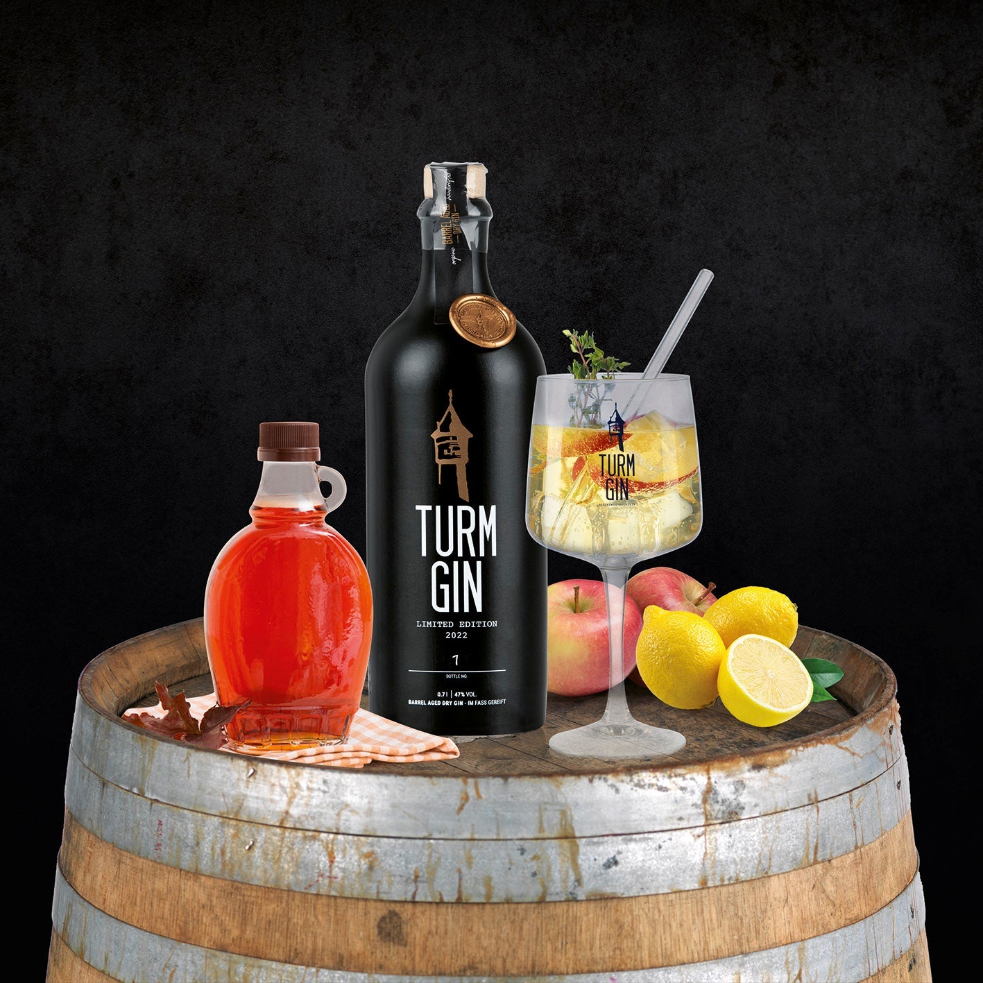 Schwarze Flasche der TURM GIN Barrel Aged - Limited Edition 20223 daneben Ahorn-Sirup in einer Flasche und ein fertiger Gin auf einem Holzfass.