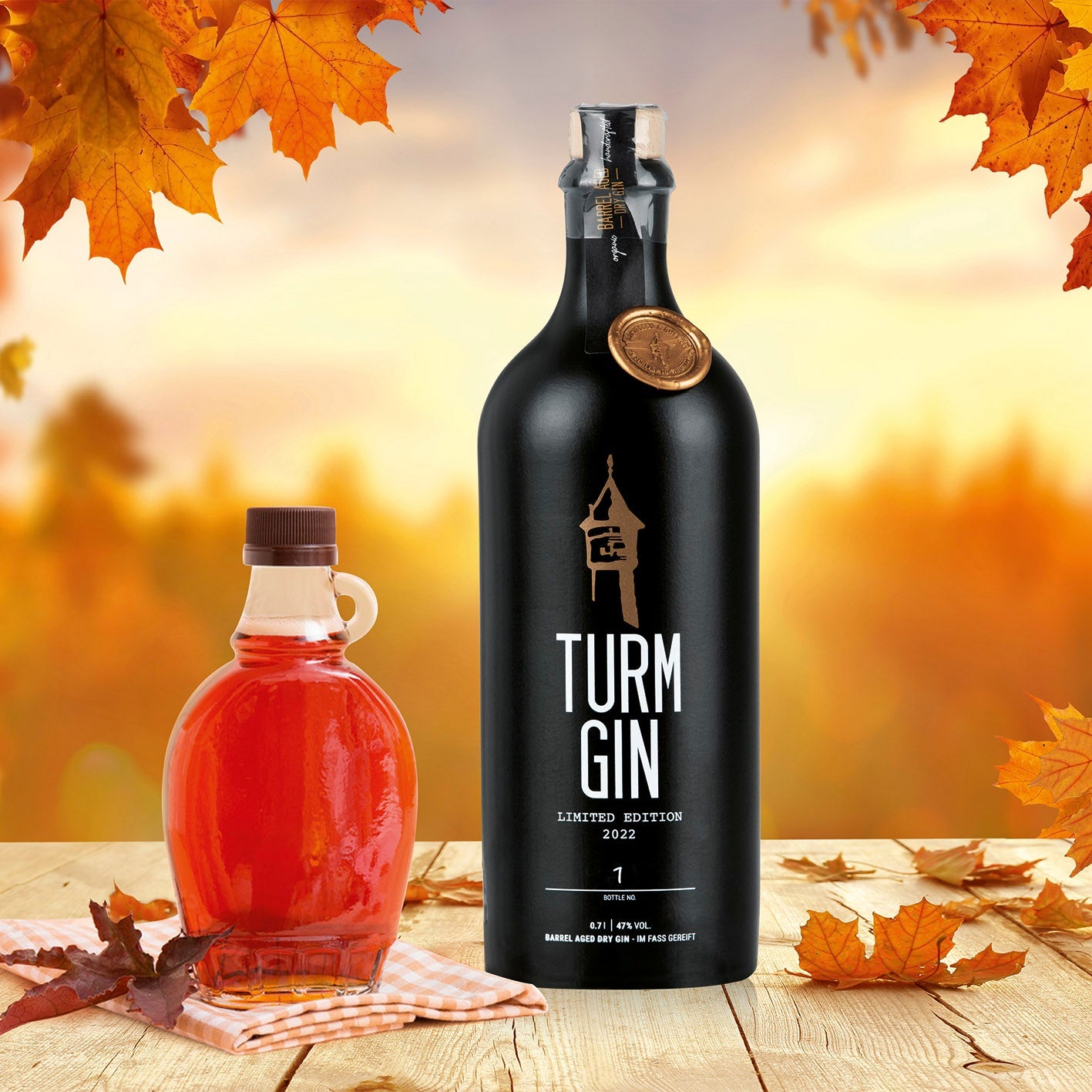 Schwarze Flasche der TURM GIN Barrel Aged - Limited Edition 20223 daneben Ahorn-Sirup auf einem Holztisch, im Hintergrund eine herbstliche Szene.