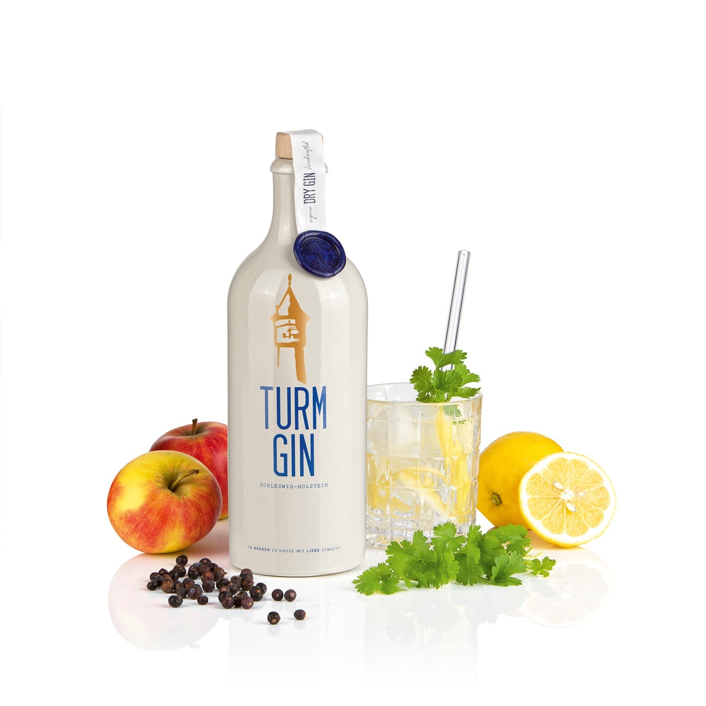 Weiße Flasche TURM GIN Dry Gin mit Turmsiegel, daneben Äpfel, Zitronen und einem Cocktailglas mit Minze vor weißem Hintergrund.