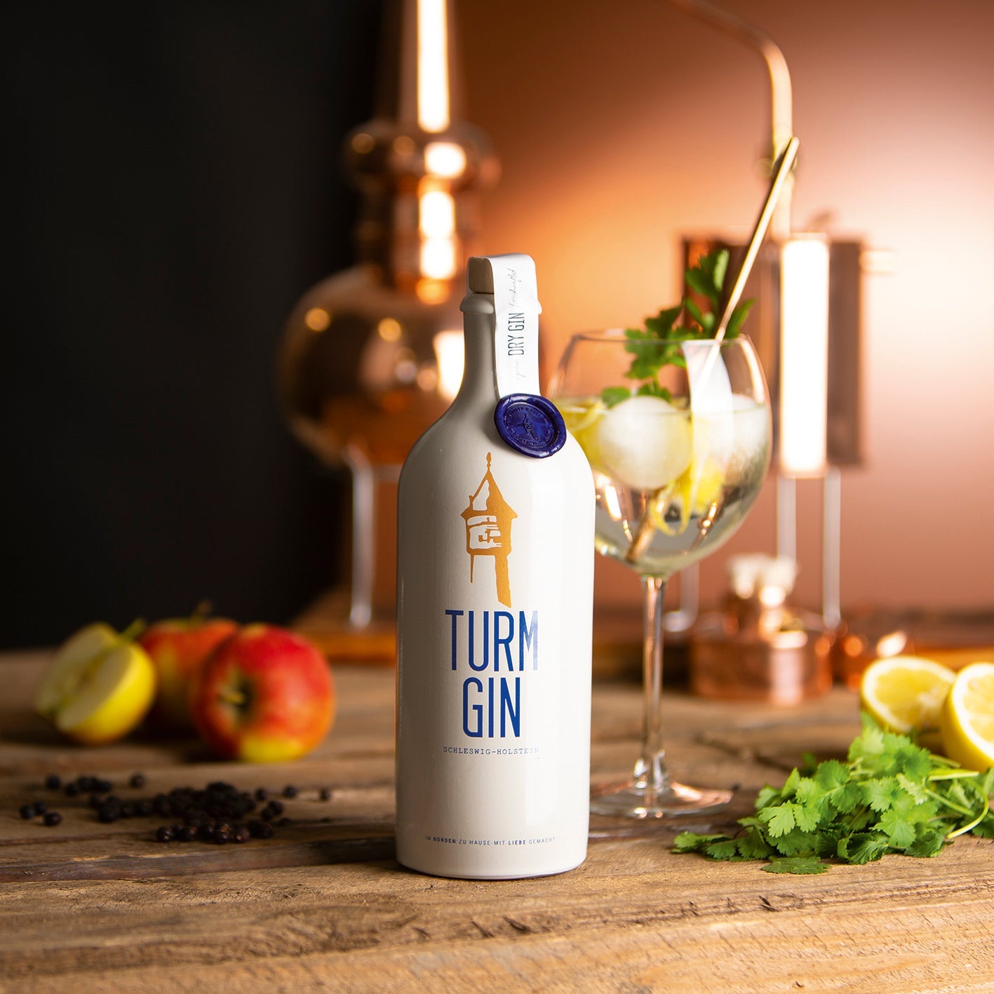 Weiße Flasche des TURM GIN Dry Gin mit Turmsiegel auf einem Holztisch, dahinter Äpfel, Minze, Zitronen und ein fertiger Cocktail.