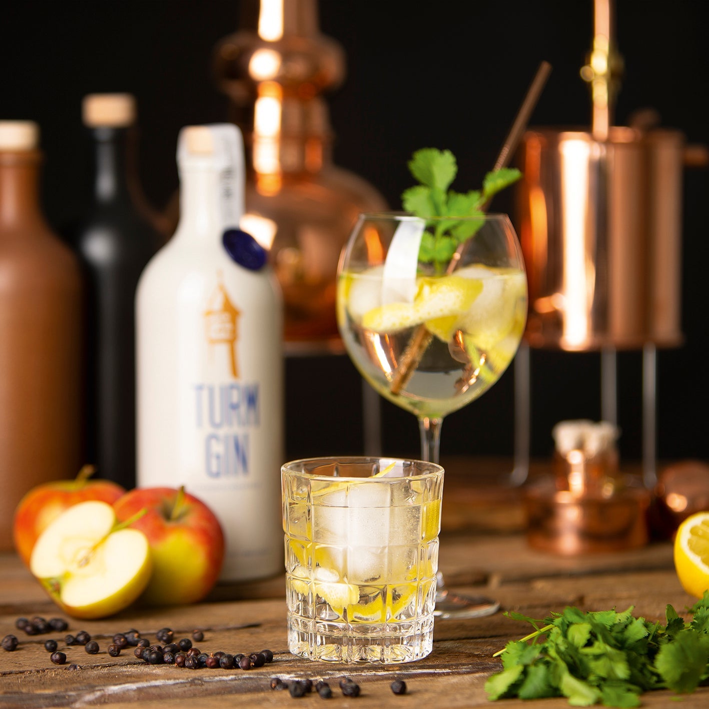 Gin in zwei unterschiedlichen Gläsern mit Zitrone und Minze, dahinter die weiße TURM GIN Flasche, Äpel und einer Destillerie.